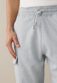 Main d'homme à côté d'un pantalon de survêtement gris clair avec ceinture élastique, cordon de serrage et rabat de poche sur la cuisse sur un fond neutre.