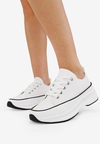 White canvas sneakers with a rounded toe, black sole, and rubber accents. Features six metal eyelets and flat laces.