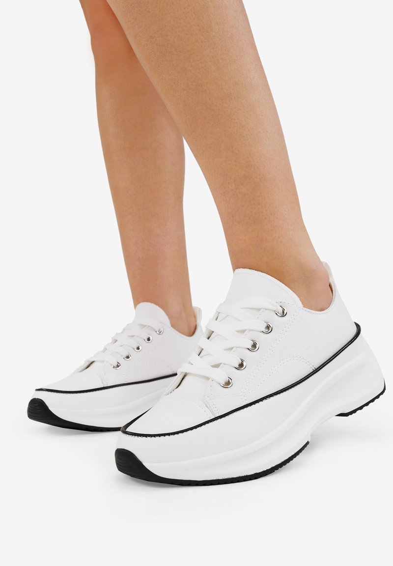 White canvas sneakers with a rounded toe, black sole, and rubber accents. Features six metal eyelets and flat laces.