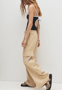 Pantaloni cargo beige con gambe ampie, vita regolabile con cordino e etichetta a patch. Top senza maniche nero con dettagli bianchi e schiena scoperta. Sandali neri.