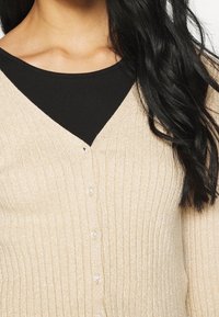 Light beige ribbed cardigan with a v-neckline, featuring clear buttons and a shimmer texture. Layered over a black top.