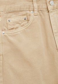 Gros plan sur la poche avant d'un pantalon en denim beige avec petite poche pour pièce de monnaie, coutures visibles, rivets et bouton au-dessus de la ceinture.