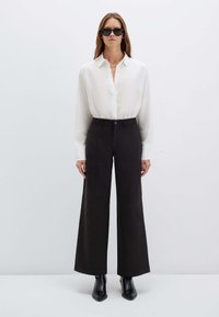 Weiße Bluse mit langen Ärmeln, kombiniert mit hochgeschnittenen, weit geschnittenen schwarzen Hosen. Schwarze Knöchelstiefel runden das Outfit ab. Minimalistisches Design, leichtes Material.