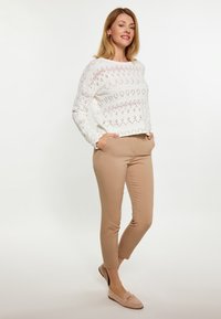 Pullover bianco lavorato a maglia con motivo a diamante, abbinato a pantaloni aderenti color sabbia e mocassini beige, che mostrano un look casual ma curato.