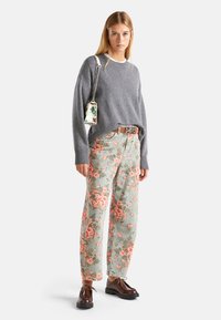 Pull en laine grise avec des poignets côtelés, pantalon à motifs floraux en vert menthe et rose, chaussures marron à lacets, et sac floral avec anse chaînes.