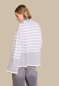 Grå og hvit stripete genser med høy krage, belledetaljer og ribbede mansjetter, sammen med lysegrå bukser, mot en nøytral bakgrunn.