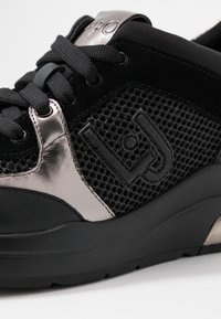 Sneaker nera con materiali in rete e suede, dettagli metallici argento, motivi testurizzati e suola in gomma con lacci neri.