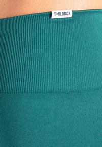 Türkisfarbener, gerippter Bund mit glatter Textur, versehen mit einem kleinen weißen Logo-Tag. Der Stoff wirkt dehnbar und körperbetont.