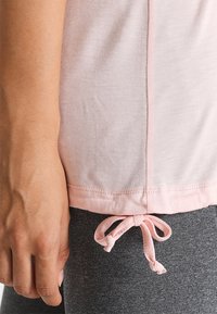 Jasnoróżowy bawełniany top z boczną kokardką przy dolnym brzegu, zestawiony z szarymi legginsami. Miękka tekstura, prosta forma.