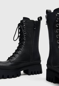Bottes de combat noires en cuir à lacets avec fermeture éclair latérale et semelles épaisses à crampons sur fond gris clair.