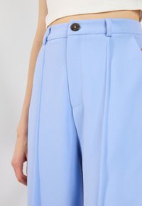 Detailní záběr na osobu v světle modrých kalhotách s vysokým pasem, černým knoflíkem a bílém tričku zasunutém.