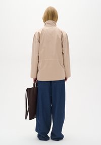 Beige voděodolná bunda s vysokým límcem, v kombinaci s volnými modrými džínovými kalhotami. Drží hnědou koženou tašku. Minimalistický design.