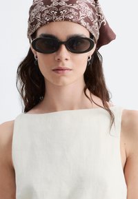 Femme portant des lunettes de soleil ovales foncées, des boucles d'oreilles argentées en créoles, un foulard marron à motifs et un haut blanc sans manches sur un fond uni.