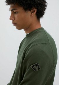 Maglione verde con collo a coste e tessuto testurizzato. Presenta una toppa triangolare sulla manica sinistra con il logo del marchio.