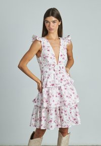 Ärmelloses Kleid mit floralem Muster, tiefem V-Ausschnitt, Rüschenschultern, gestuftem Rock und einem Gürtel mit einer runden Schnalle. Heller rosa Hintergrund.