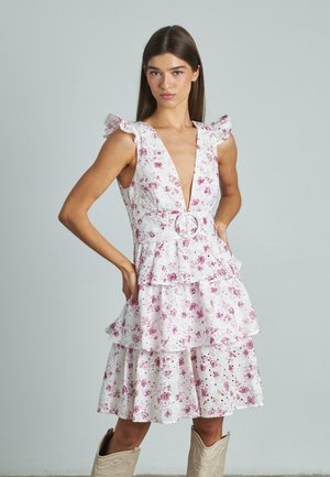 Ärmelloses Kleid mit floralem Muster, tiefem V-Ausschnitt, Rüschenschultern, gestuftem Rock und einem Gürtel mit einer runden Schnalle. Heller rosa Hintergrund.