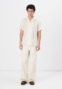 Helles beige Kurzarmhemd mit floraler Stickerei und Knopfleiste, kombiniert mit crema-farbenen, weiten Hosen und dunklen braunen Schuhen.