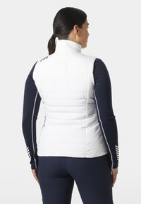 Mujer con cabello oscuro recogido en una coleta, vistiendo un chaleco acolchado blanco sobre una camiseta azul marino de manga larga con detalles de rayas en las mangas, vista desde atrás.