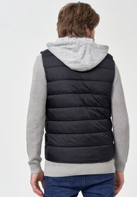 Ærmeløs sort puffervest med et gråt strikbaselag og en grå hætte. Har horisontale syninger og ribbede manchetter for tekstur.