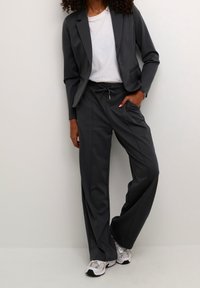 Blazer gris a medida con pantalones de pierna ancha a juego; hecho de una tela suave con un brillo sutil. Camiseta blanca debajo, llevada con zapatillas.