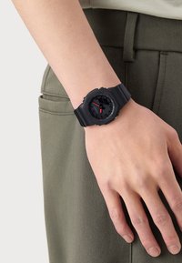 Montre numérique noire avec un boîtier rond, un bracelet en silicone, des accents rouges sur le cadran, des boutons texturés et un design minimaliste.