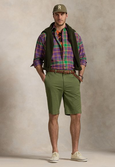 Modelo com camisa de padrão xadrez nas cores roxo, verde e azul, calções verde-oliva e ténis claros, com uma camisola pendurada nos ombros.