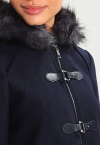 Marinblå ullkappa med huva i fuskpäls, silverfärgad dragkedja och läderdetaljer med knäppning med knappar. Mjuk textur och skräddarsydd passform.