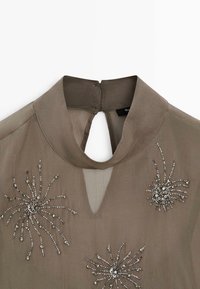 Brun gennemsigtig bluse med høj krave og nøglehuldetalje, pyntet med indviklede perledekorationer i sølv- og klare toner.