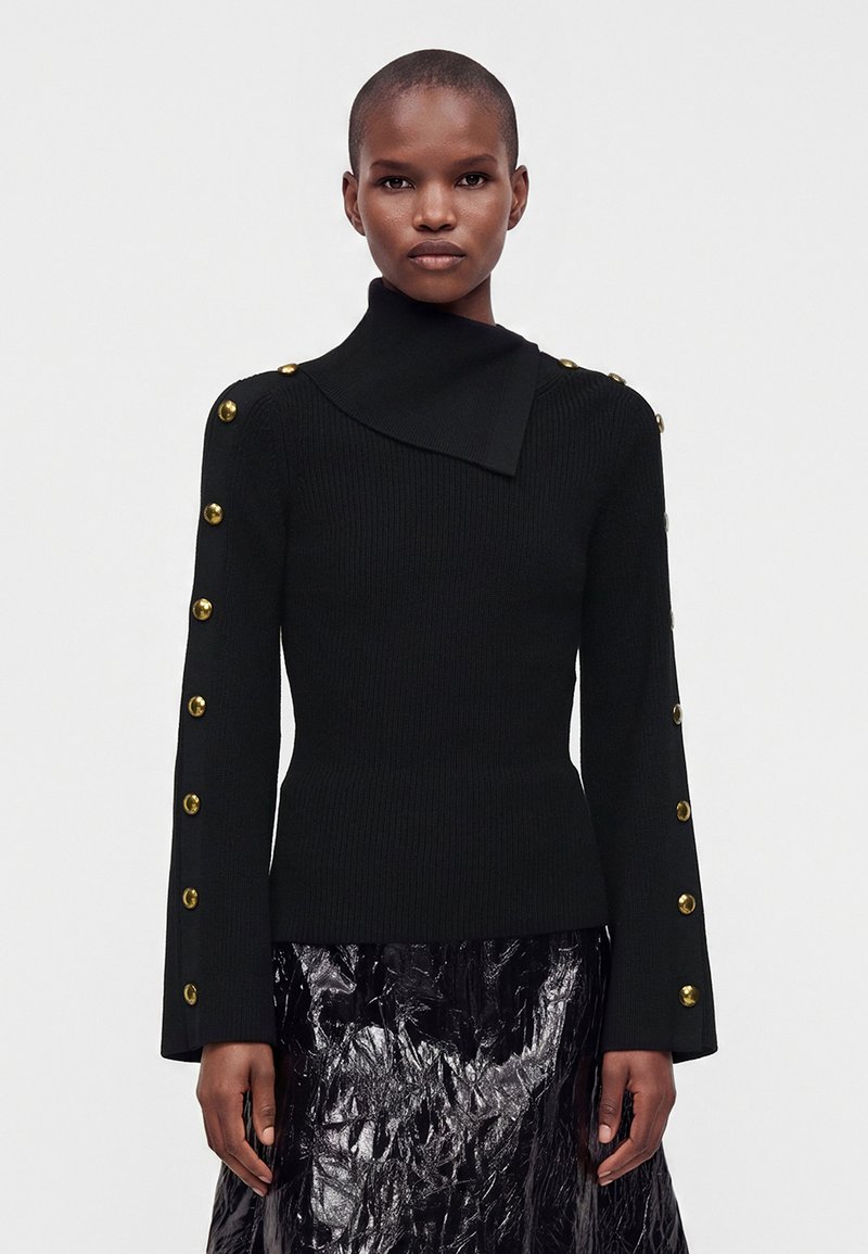 Femme portant un pull noir côtelé avec des boutons dorés le long des manches et un col haut asymétrique, associé à une jupe noire brillante.