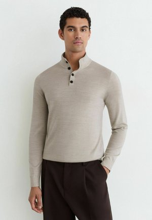 Man draagt een beige lange mouwen trui met een hoge kraag en zwarte knopen, gecombineerd met een donkere broek, staand tegen een witte achtergrond.