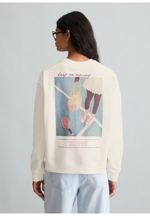 Femme aux longs cheveux foncés portant des lunettes et un sweat-shirt blanc avec un motif sur le thème du tennis et le texte « keep on moving. »