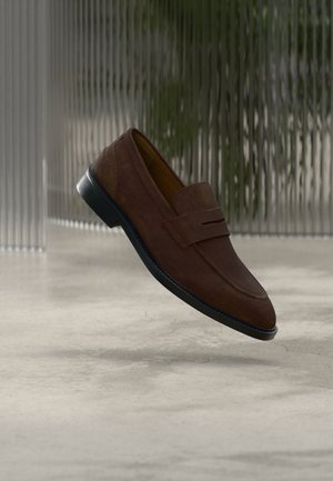 Bruine suède loafers met een gladde textuur, een lichte hak en een opvallende decoratieve riem aan de voorkant. Zwarte rubberen zool.