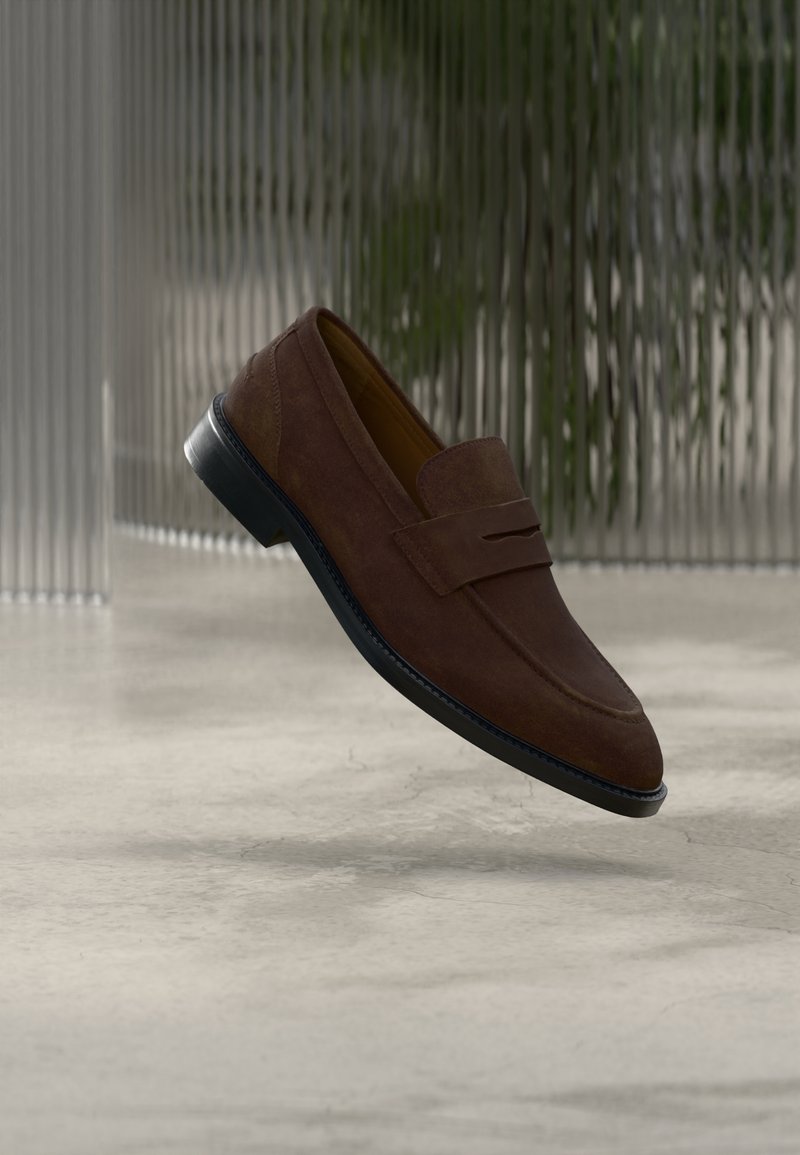 Καφέ μοκασίνια από σουέντ με απαλό ύφασμα, ελαφριά σόλα με τακούνι και ένα εμφανές διακοσμητικό λουράκι στο μπροστινό μέρος. Μαύρη λαστιχένια σόλα.