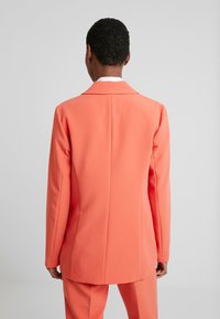 Koralowa marynarka w gładkim materiale o dopasowanym kroju, z klapami z wycięciem, pojedynczym rozcięciem z tyłu i długimi rękawami.