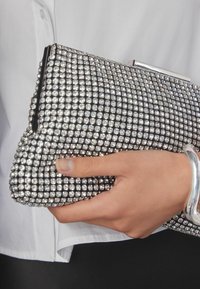 Hand die een kleine zilveren clutch met strass-steentjes vasthoudt, gekleed in een wit overhemd en een glanzende zilveren armband draagt.
