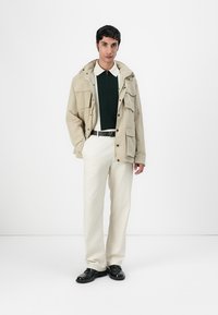 Veste beige avec plusieurs poches, polo vert, pantalon crème et chaussures noires. La veste a une capuche et des boutons à pression.
