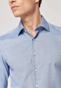 Camicia button-up azzurro chiaro con un motivo a micro-check, colletto a punta e bottoni bianchi rotondi. Il tessuto strutturato crea una finitura liscia.