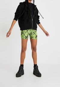 Sudadera negra con cremallera, pantalones cortos de estampado de serpiente verde neón y botas negras gruesas con plataforma. El conjunto presenta un audaz contraste de colores y patrones texturizados.