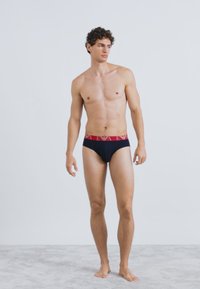 Cuecas de algodão pretas com cintura vermelha, apresentando detalhes de logotipo branco, ajuste justo e textura suave. Modelo está em pé contra um fundo neutro.