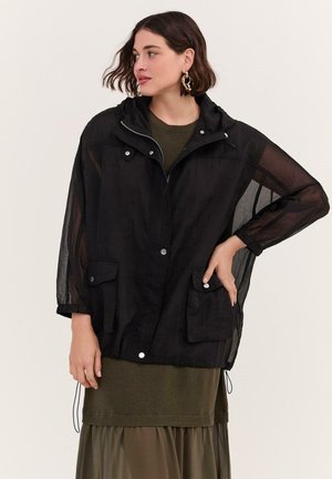 Kvinde iført gennemsigtig sort hættetrøje med forlommer over olivengrønt strikket kjole, kigger til venstre med hånden på hoften.