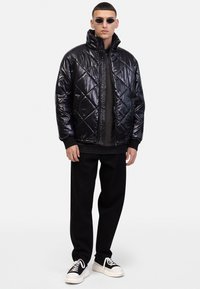 Zwarte, gewatteerde jas met ruitpatroon, hoge kraag en glanzende textuur. Draagt over een donkere top en gecombineerd met losse zwarte broeken en sneakers.