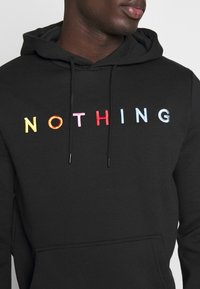 Sudadera negra con la palabra "NADA" bordada en multicolores. Tiene capucha con cordón y un bolsillo frontal. Tejido de textura suave.