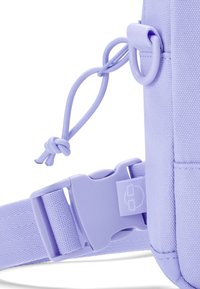 Lila nylonväska med texturerat tyg, justerbar rem, plastspänne och snördetaill. Har en logotyp på spännet.