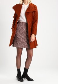 Cappotto color ruggine con grande collare, dettagli in bottoni, abbinato a una gonna testurizzata di lunghezza al ginocchio. Calze di rete nere e scarpe lucide completano il look.