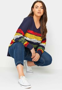 Laivastonsininen pitkähiainen paita, jossa on monivärinen vaakasuora raidallinen tekstiuri. Yhdistetty sinisiin revittyihin farkkuihin ja valkoisiin kangaskenkiin.