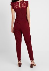 Bordeaux kombinezon z koronkową górą, z wycięciem na plecach i falbankowymi rękawami. Rozszerzane nogawki i dopasowany pas, stylizowany z sandałami na obcasie w kształcie bloku.