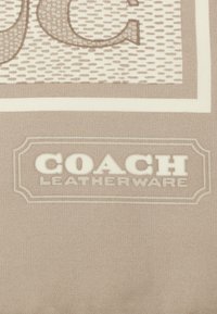 Beżowy materia�ł z teksturowanym wzorem i białym logo "COACH LEATHERWARE". Ozdobna ramka otacza logo.