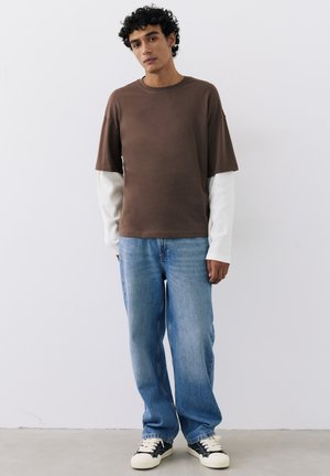 Jonge man met krullend haar die een gelaagd bruin-wit shirt met lange mouwen draagt, losse blauwe spijkerbroek en zwart-witte sneakers, staand tegen een witte muur.