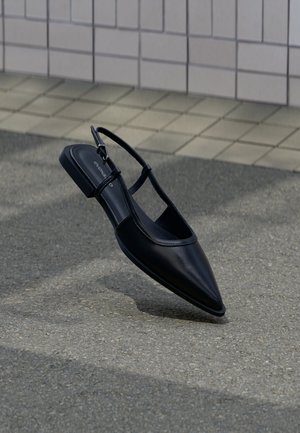 Chaussure plate noire à bout pointu avec bride arrière, flottant au-dessus d'une surface en bitume à côté d'un trottoir carrelé.