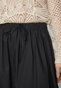 Svart, texturerad tygkjol med elastisk midja och en knytbar dragsko-detalj framtill, delvis lagd över en vit virkatröja.